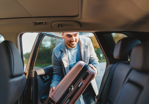 Man placing a travel suitcase in the back seat of his rental car before heading to a business event.