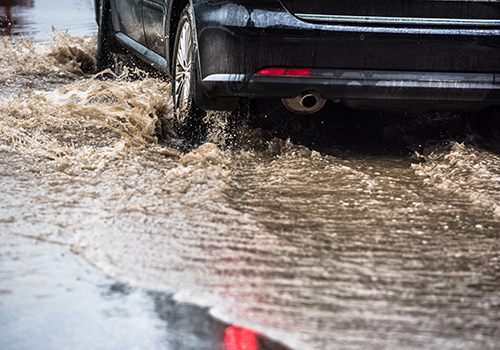 Car wheel driving through a flooded street, with water splashes showing risk of aquaplaning during heavy rain.