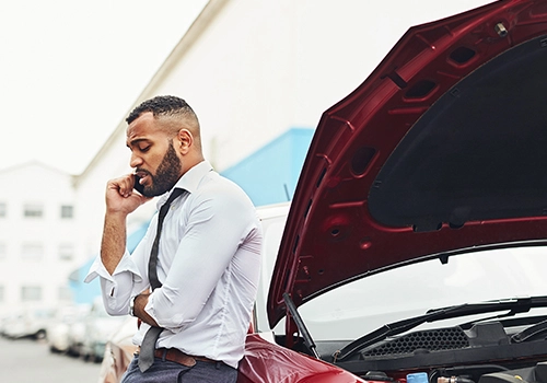 Empresário encostado a um carro vermelho com capot aberto, a falar ao telemóvel após uma avaria na via pública.