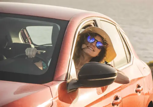 Mulher de chapéu e óculos de sol sorri, com a cabeça de fora da janela do carro