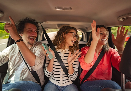 Three people sitting inside a car, laughing and singing during a trip.