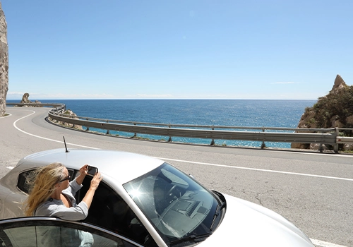 Driver outside a parked car on a scenic coastal road taking a photo of the seaside landscape during a road trip.