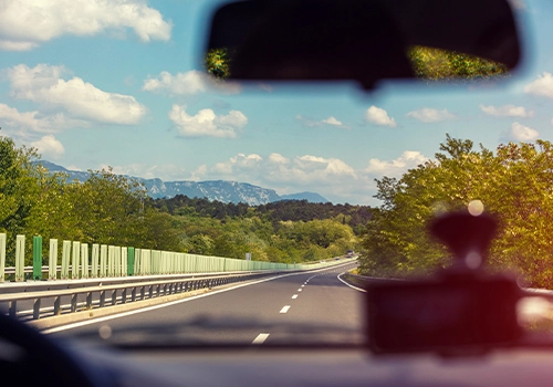 View of the road from inside a car on a motorway, with green landscape and mountains in the background.