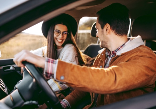 Couple inside a car on a road trip, smiling at each other, with the window open and a rural landscape in the background.