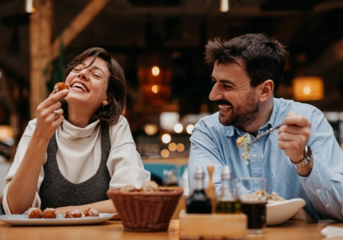 Woman and man dining in a cozy restaurant, laughing while sharing food and drinks at a warmly lit table.