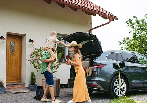 Family outside their home loading luggage into the car boot, with one adult lifting a child, getting ready for a family road trip.