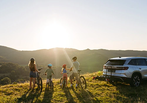 Family with children on bicycles in a meadow at sunset, with a parked car and a view of the mountains.