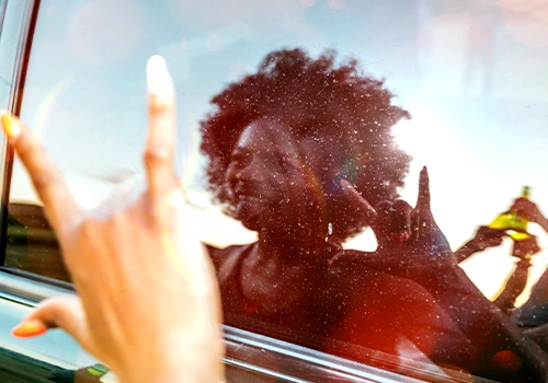Festivaleira junto a um carro, refletida no vidro, a sorrir e a fazer gesto do